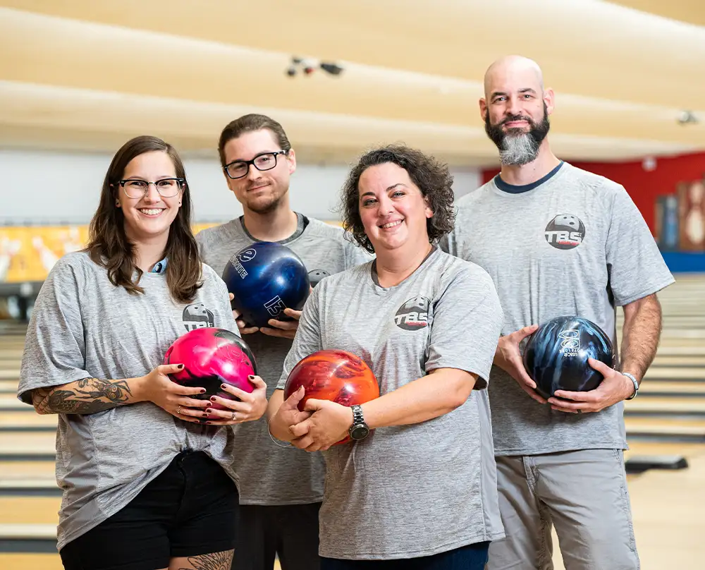 Group of four bowlers in matching gray team shirts holding colorful bowling balls at Bel-Air Bowl in Belleville, IL - Adult Bowling Leagues