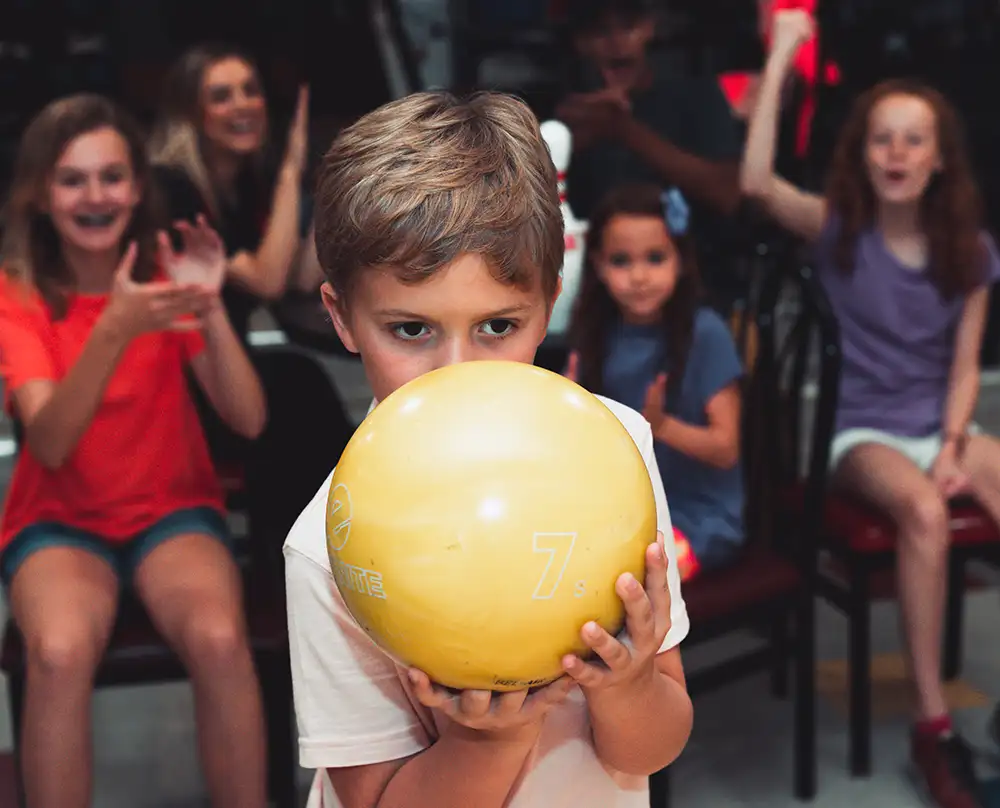 Young boy holding a yellow bowling ball up to his face while kids cheer behind him at Bel-Air Bowl in Belleville, IL