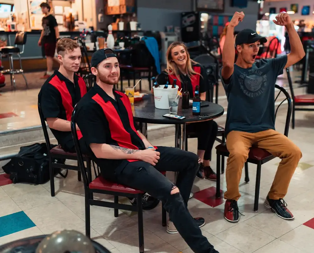 Bowlers in red and black retro shirts seated at a table while a friend cheers at Bel-Air Bowl in Belleville, IL - join a bowling league