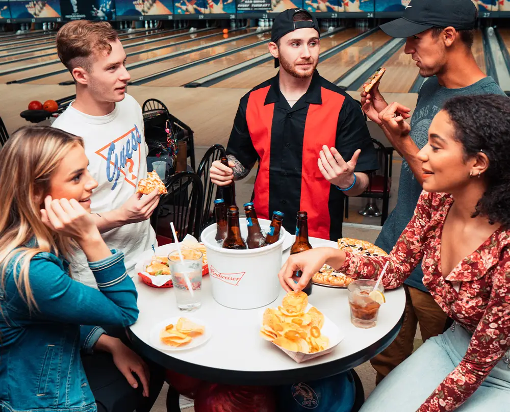 Friends sharing pizza, chips, and beer around a table with bowling lanes behind them at Bel-Air Bowl in Belleville, IL - Team Building Events
