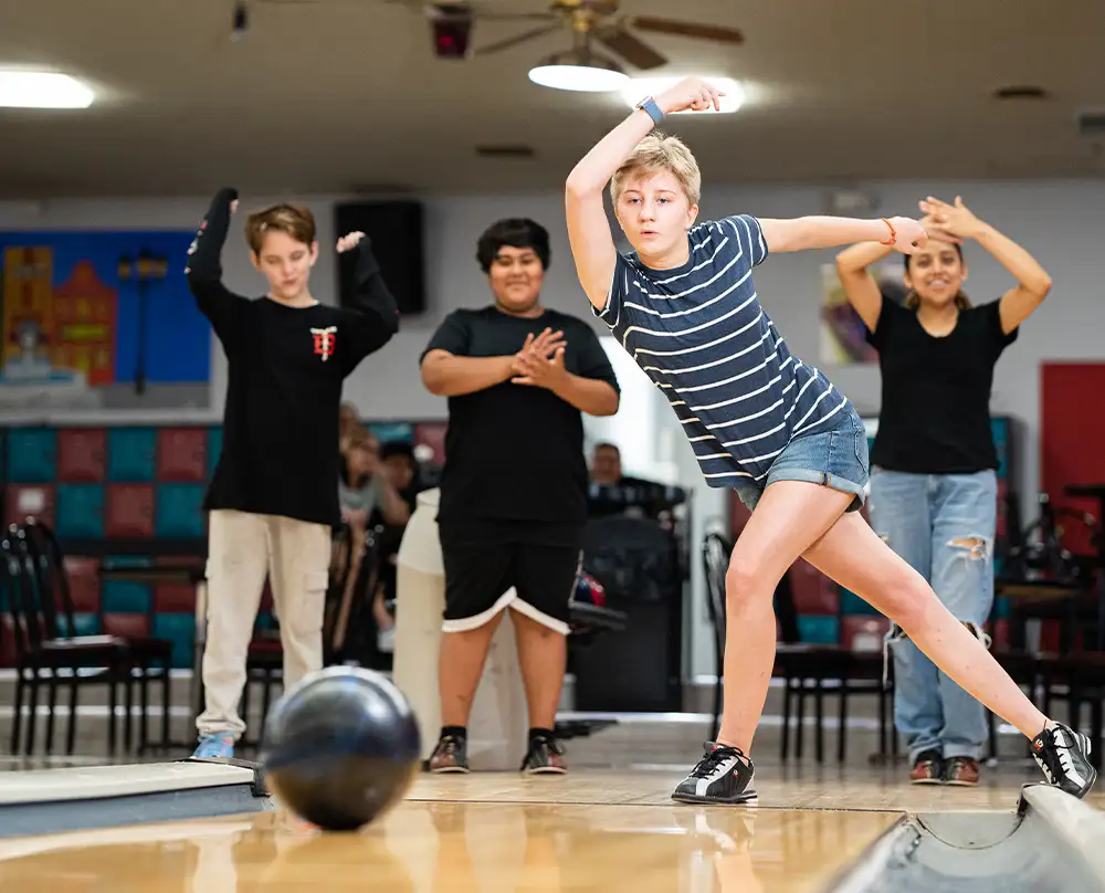Teen mid-throw on the approach as friends in Youth Bowling League cheer behind her at Bel-Air Bowl in Belleville, IL