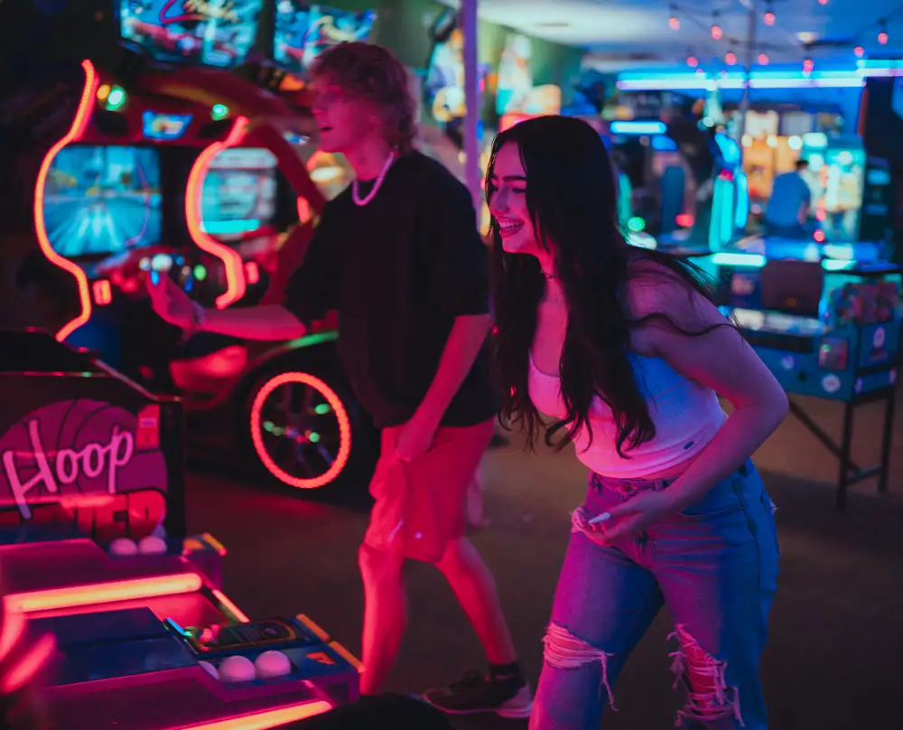 Two friends laughing at neon-lit arcade games and basketball hoops at Bel-Air Bowl in Belleville, IL